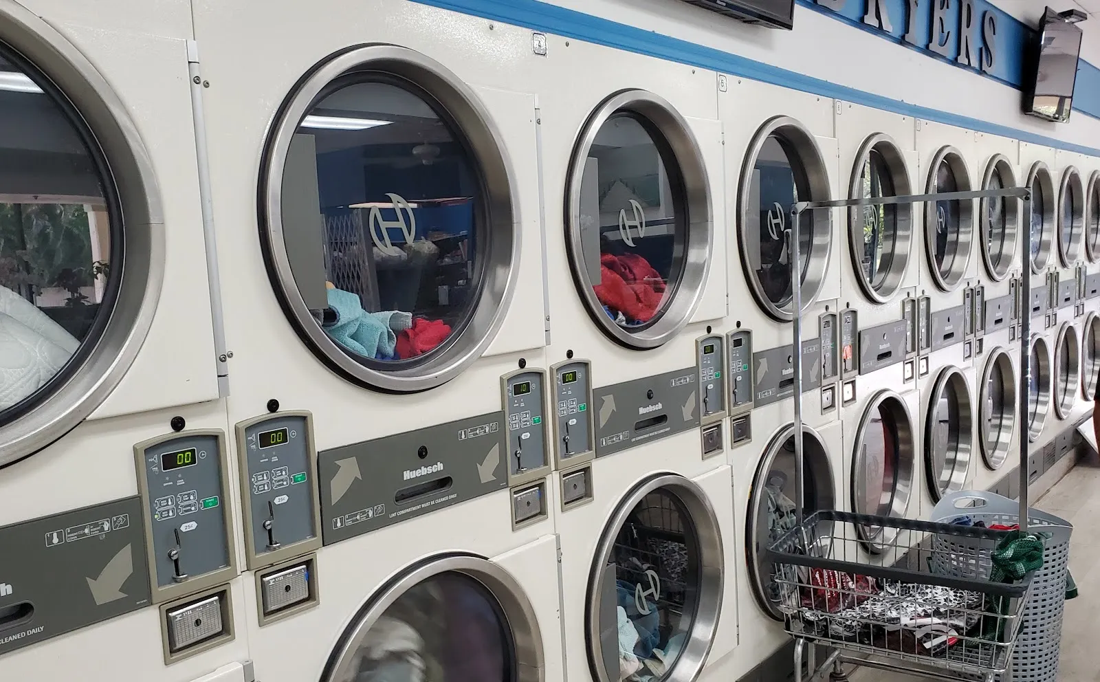 Row of stacked commercial dryers in use, with a 'Dryers' sign mounted above and laundry visible tumbling inside several machines.
